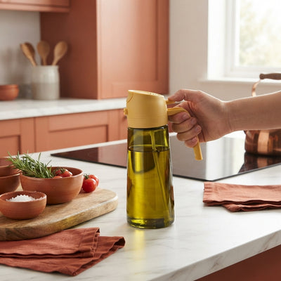 A dual-function dark glass oil spray bottle on a marble countertop with terracotta and burnt sienna kitchen accessories.